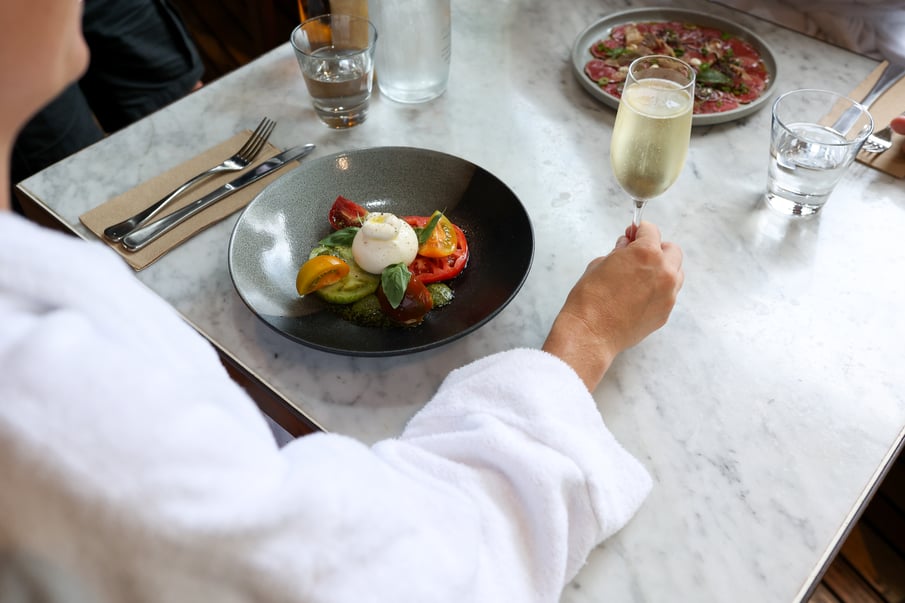 A person in a white robe drinking sparkling wine and eating a Caprese salad